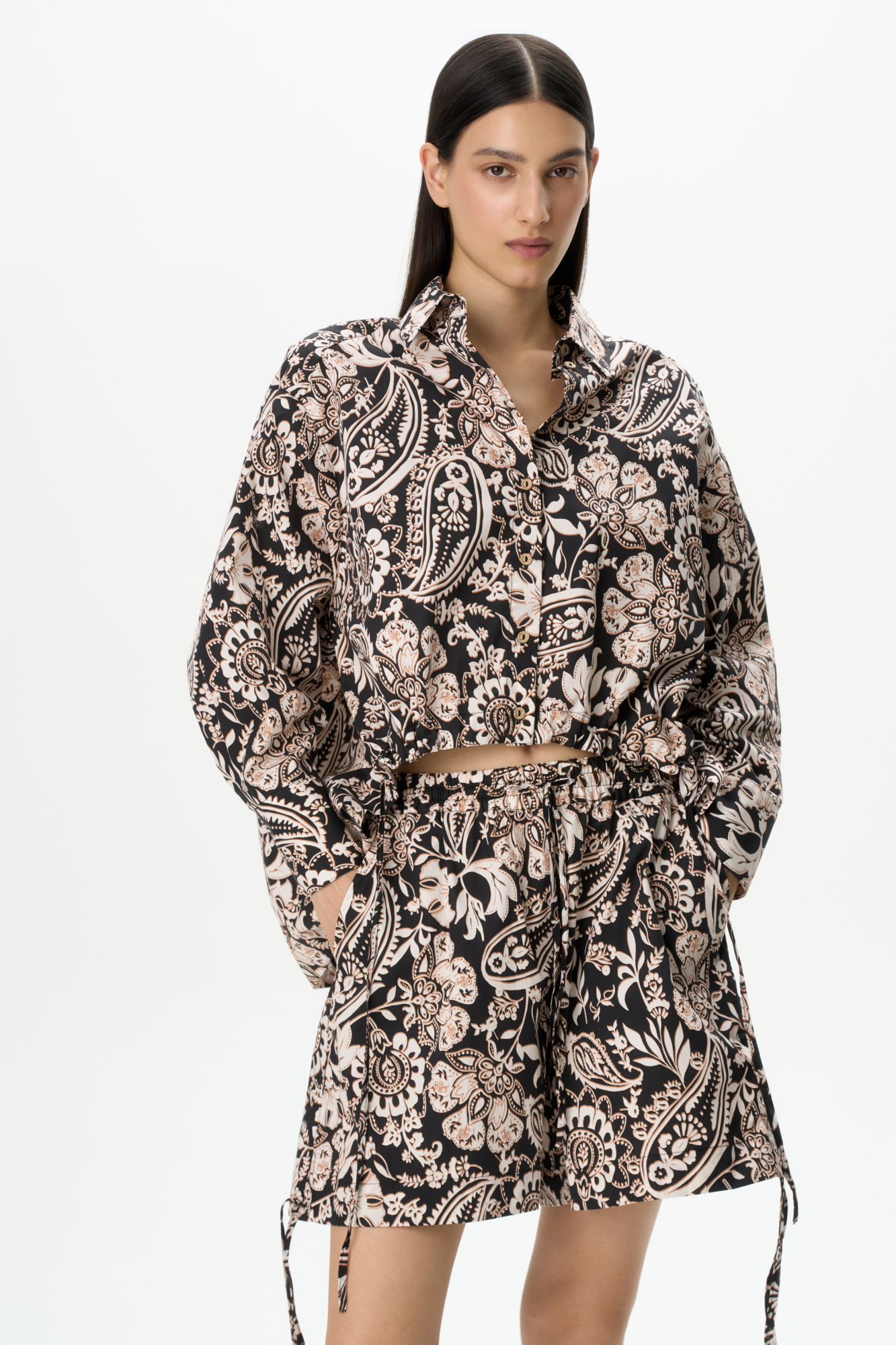 Принтованный костюм из 100% хлопка, состоящий из рубашки и шорт. Рубашка свободного кроя, с длинными рукавами, классическим отложным воротником и кулиской по низу изделия. Короткие шорты с карманами, широким поясом на кулиске и завязками по бокам.