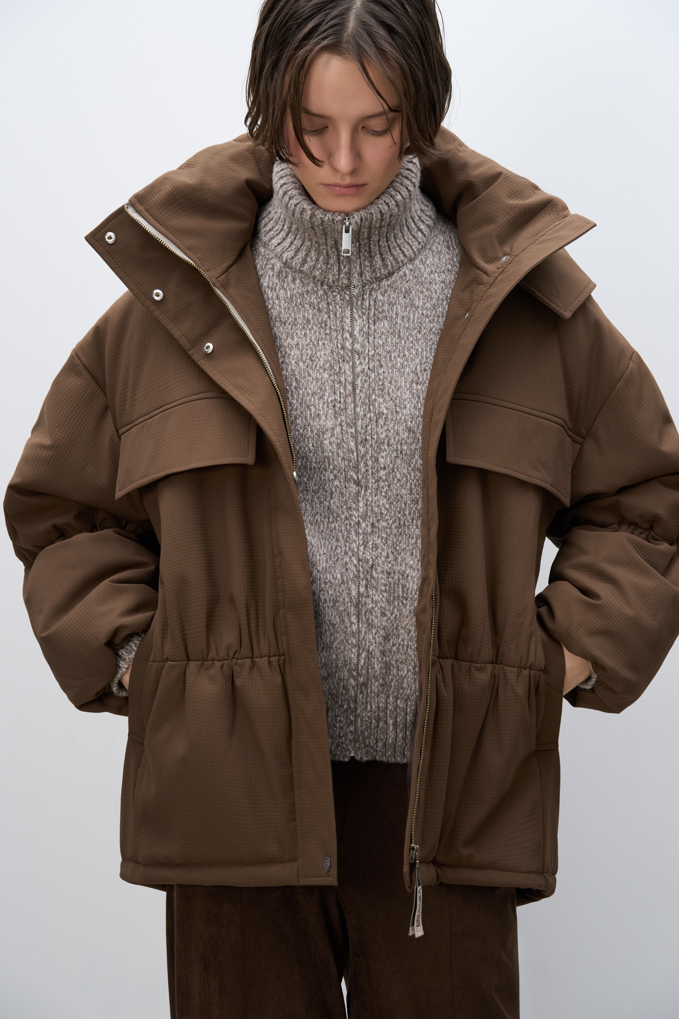 Длинная куртка на синтепоне из текстурной ткани. Модель свободного силуэта со спущенной линией плеча, накладными и боковыми карманами, воротником-стойкой и капюшоном. Изделие на подкладке. Застежка на молнию под планкой с кнопками.