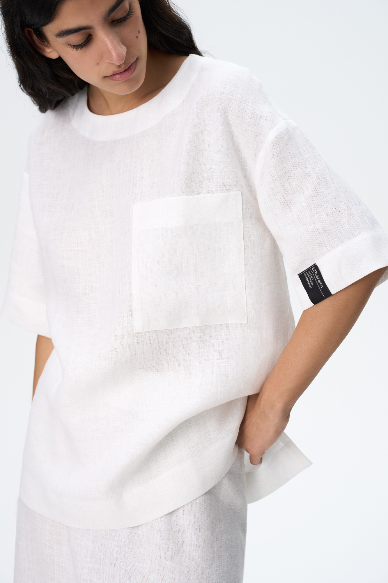 Широкая футболка из 100% льна. Модель прямого кроя, со спущенной линией плеча, круглым вырезом горловины и асимметричным низом с разрезами по бокам. Изделие декорировано накладным карманом на груди и брендированным ярлыком на рукаве. Застежка на пуговицу сзади.