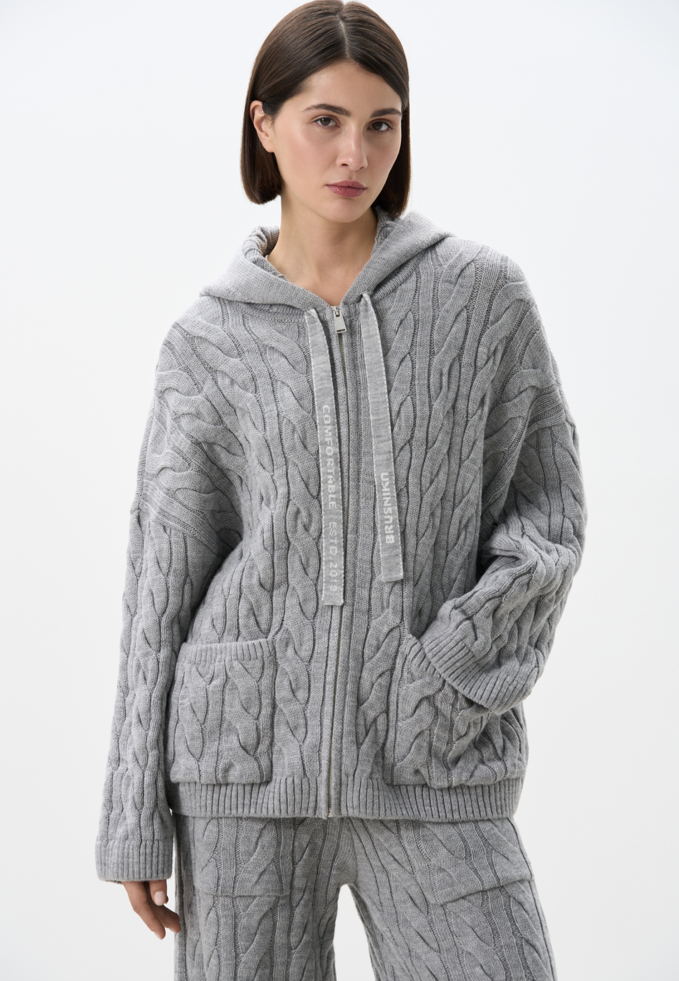 Трикотажное худи с объемными косами из мягкой шерсти. Модель свободного силуэта, со спущенной линией плеч, накладными карманами и капюшоном. Изделие декорировано брендированным жаккардом на спине. Застежка на молнию. Выполнено без подкладки. Дополнить образ можно брюками или юбкой в том же стиле.