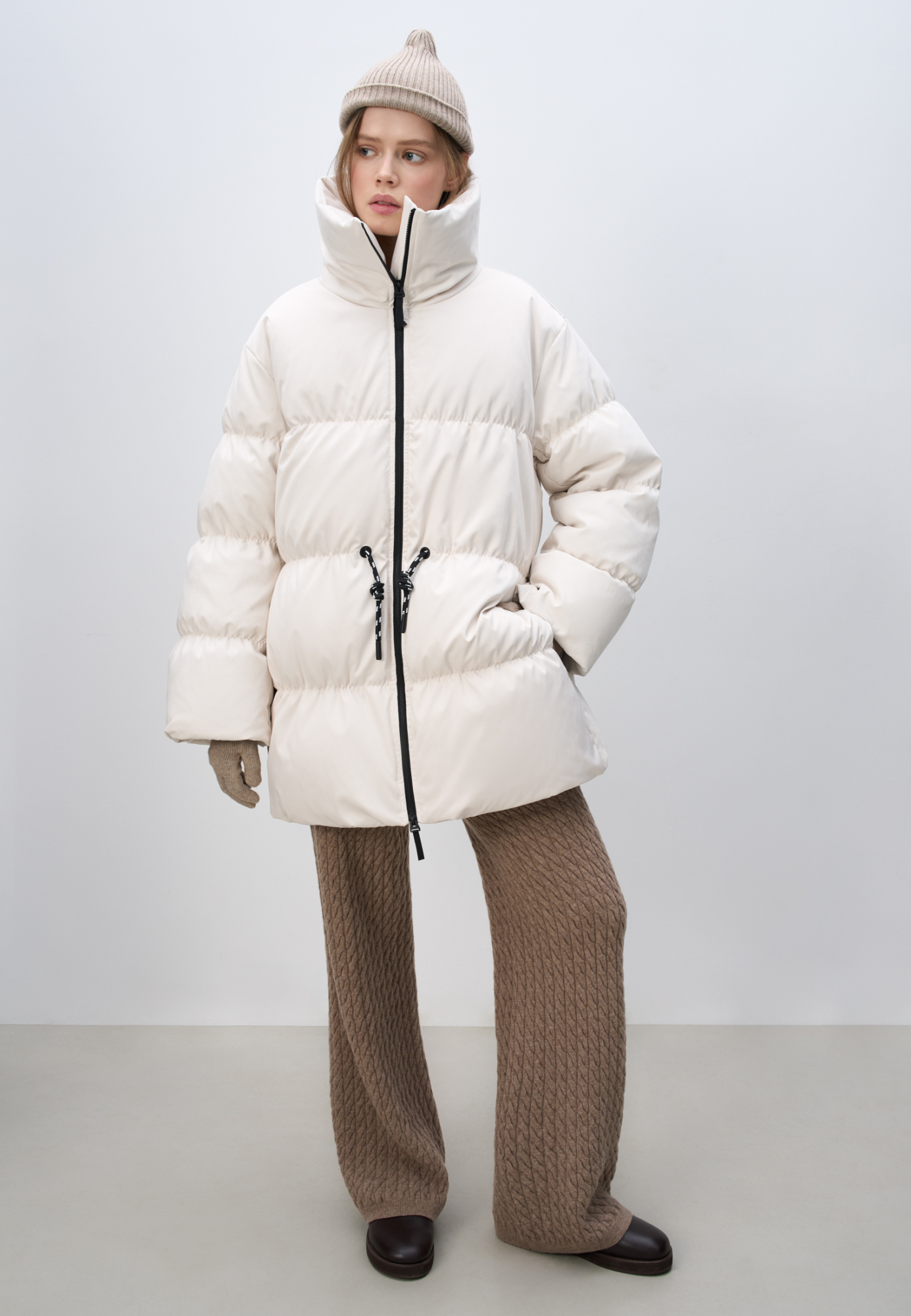 Стеганый объемный пуховик. Модель свободного силуэта с воротником-стойкой, кулиской на талии и логотипом бренда на спине. Застежка на молнию.
