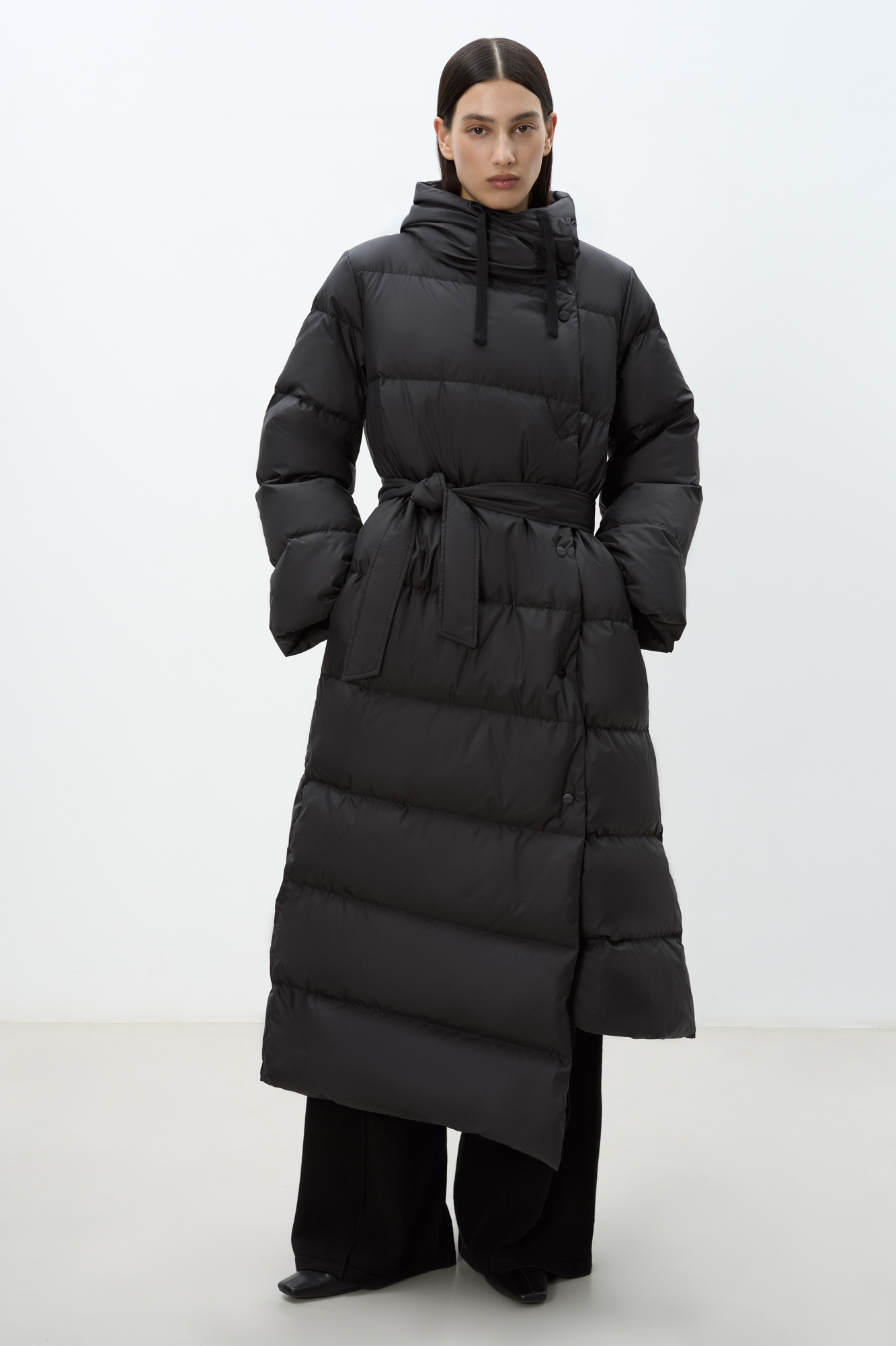 Длинный стеганый пуховик с косой застежкой на кнопки. Модель с ассиметричным низом, боковыми карманами и капюшоном. В комплекте пояс в тон.