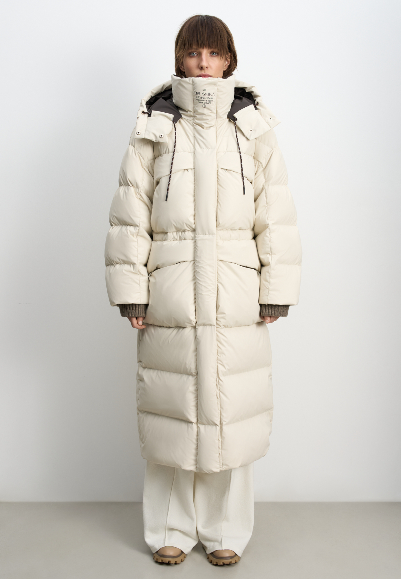 Объемный длинный пуховик. Модель с воротником-стойкой, капюшоном на кулиске и кнопках, большими нагрудными и боковыми карманами на молнии с клапанами и трикотажными манжетами в широкий рубчик. Застежка на молнию по всей длине под планкой на кнопках. Ворот изделия декорирован логотипом бренда.