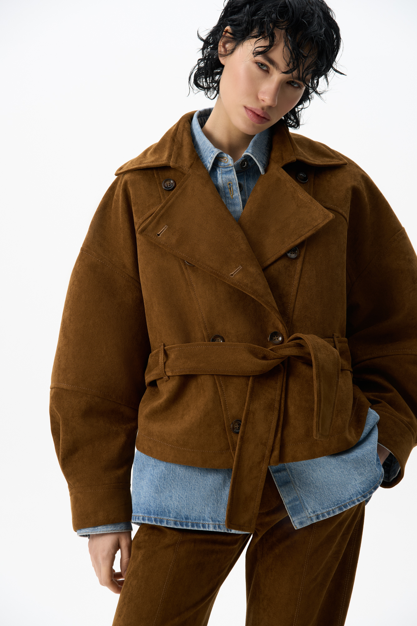 Двубортный короткий тренч сложной геометрии, выполненный из ткани с имитацией внешнего вида и свойств натуральной замши. Модель с отложным воротником, свободными рукавами со спущенной линией плеч, шлевками на поясе и ремнем.