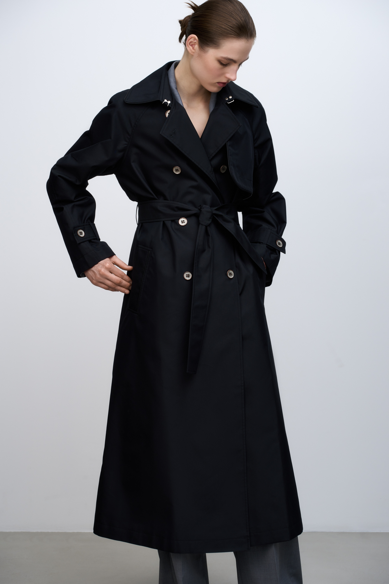 Двубортный длинный тренч прямого кроя. Модель с отложным воротником, декорированным металлической перемычкой, отлетной кокеткой, патами с пуговицами на манжетах, карманами и шлевками на талии для пояса.