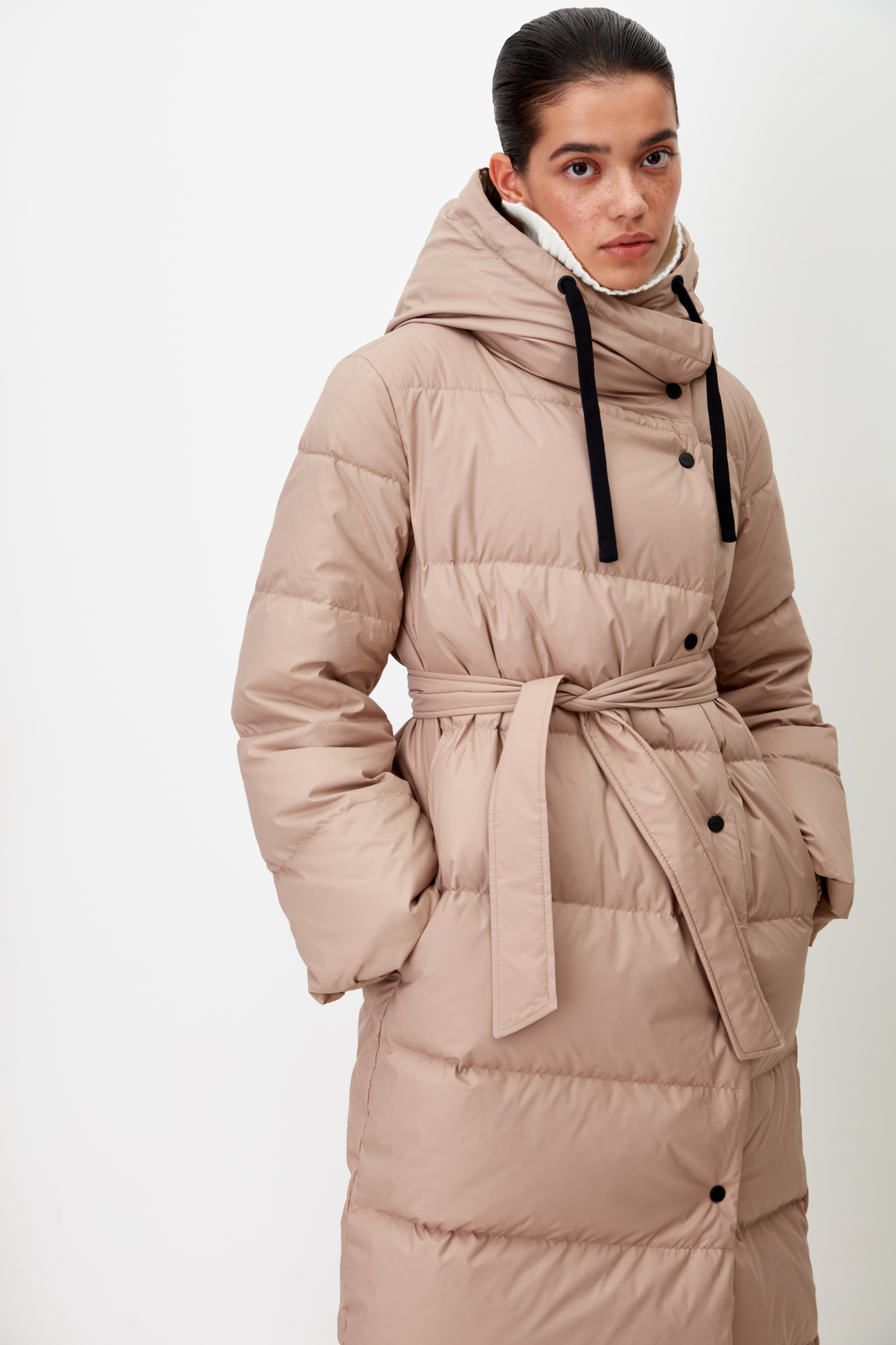 Длинный стеганый пуховик с косой застежкой на кнопки. Модель с ассиметричным низом, боковыми карманами и капюшоном. В комплекте пояс в тон.