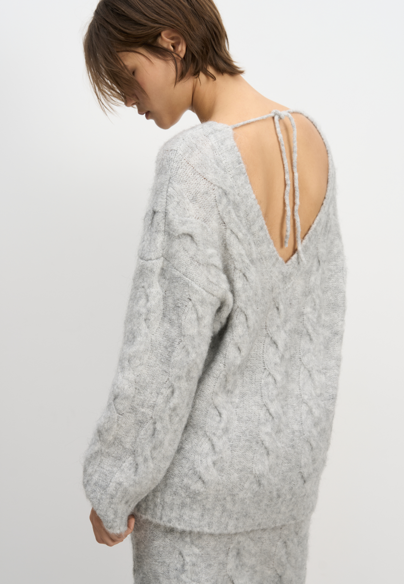 Трикотажный джемпер из шерсти и мохера. Модель свободного силуэта со спущенной линией плеч, длинными рукавами, круглой горловиной и глубоким вырезом на спине, фиксирующимся шнурком. Изделие декорировано фактурной вязкой в виде кос. Края изделия выполнены в рубчик.