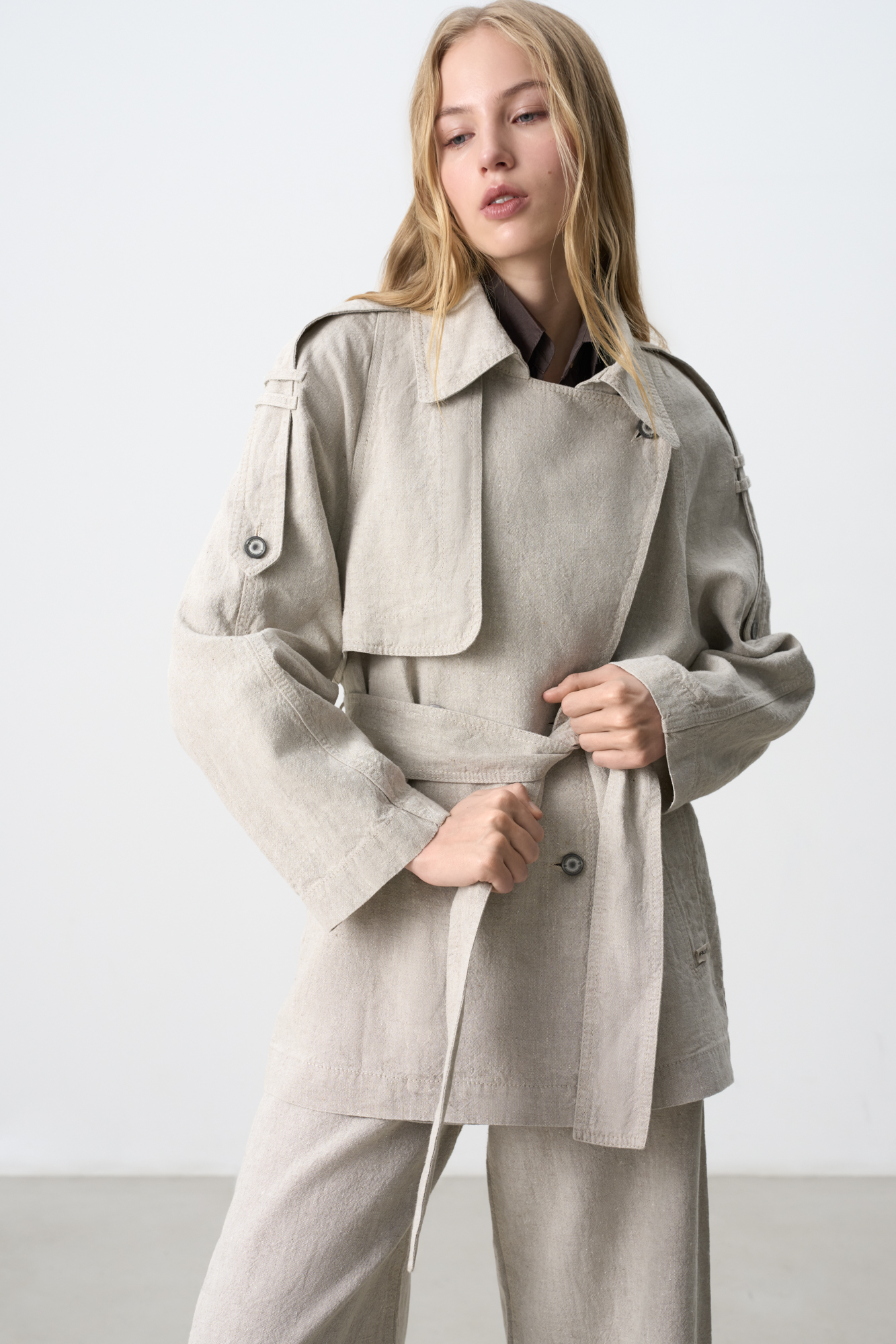 Двубортный тренч из 100% льна. Модель прямого кроя, с отложным воротником и кокеткой. Изделие декорировано патами на плечах. Два боковых кармана. Застежка на пуговицы. Пояс в комплекте. 