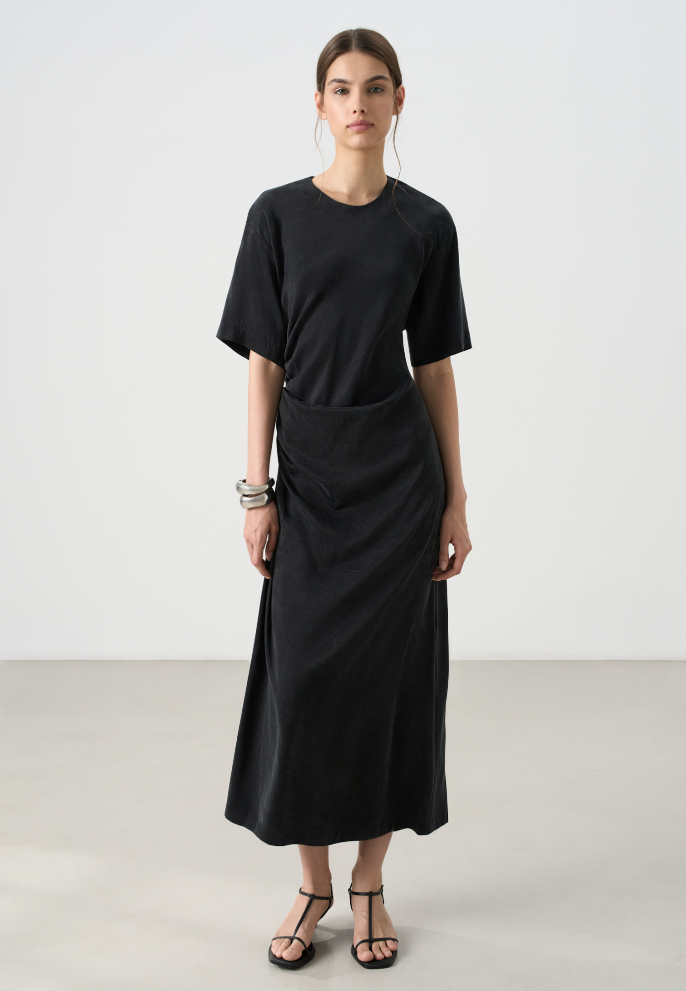 Платье длины миди из купры. Модель свободного кроя, с круглым вырезом горловины, спущенной линией плеч, короткими рукавами и драпировкой на талии сбоку. Застежка на пуговицу сзади.