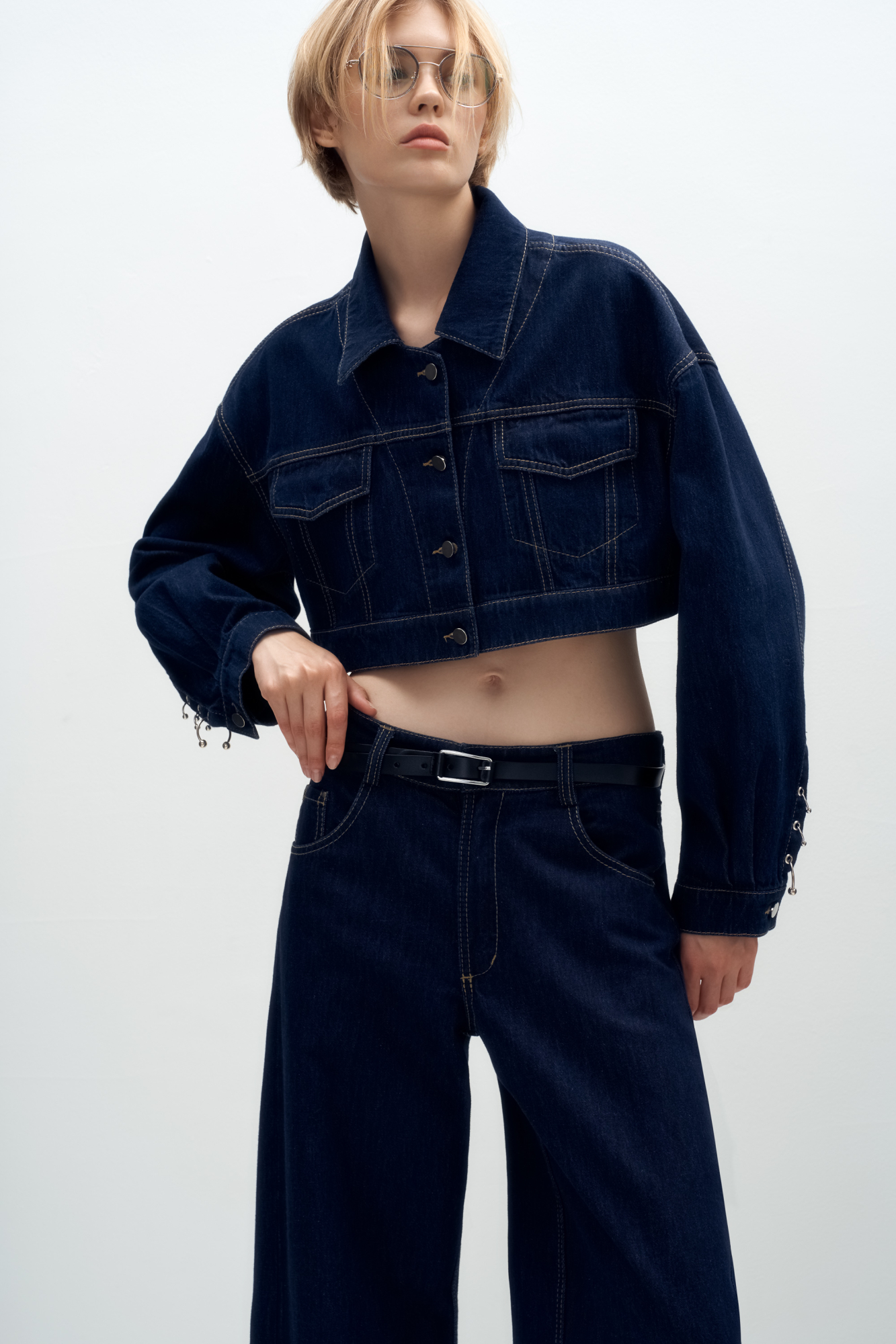 Укороченная куртка прямого кроя из денима с отложным воротником и хлястиками на поясе. Модель декорирована накладными карманами на груди, металлической фурнитурой в виде пирсинга на манжетах и брендированным ярлыком на спине. Застежка на пуговицы.
