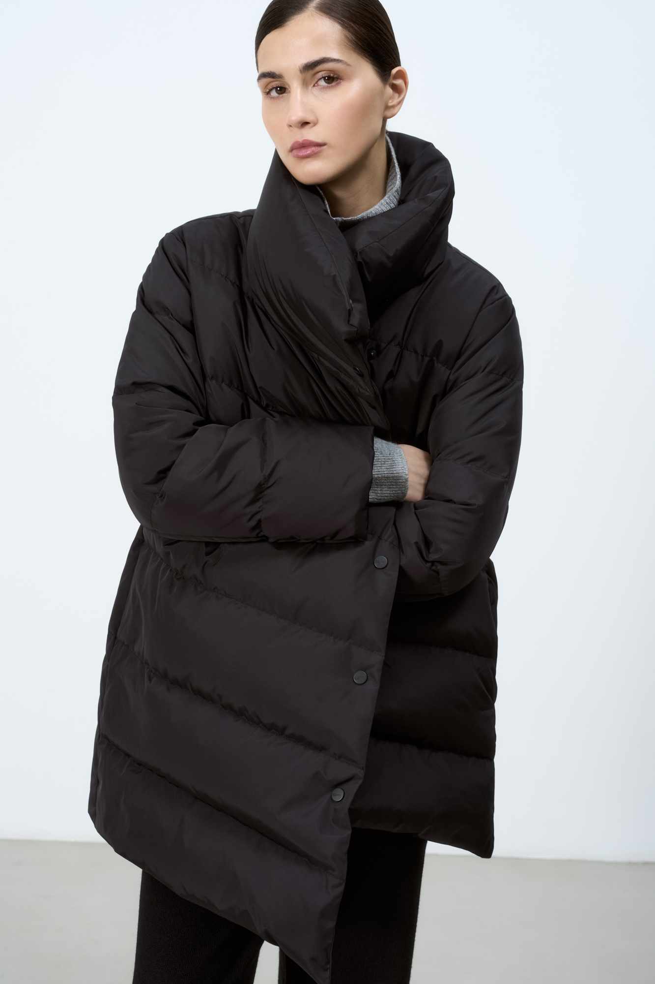 Утепленный пуховик с косой застежкой на кнопки. Модель свободного силуэта с боковыми карманами и высоким воротом. На подкладке.