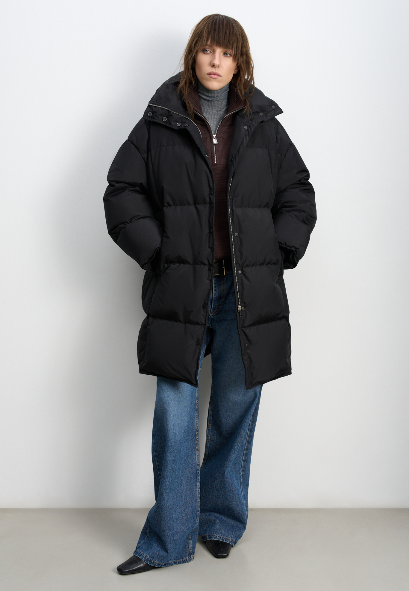 Длинный стеганый пуховик с капюшоном на кулиске. Модель свободного силуэта с воротником-стойкой, трикотажными манжетами и боковыми карманами. Застежка на молнию под планкой с брендированными пуговицами.