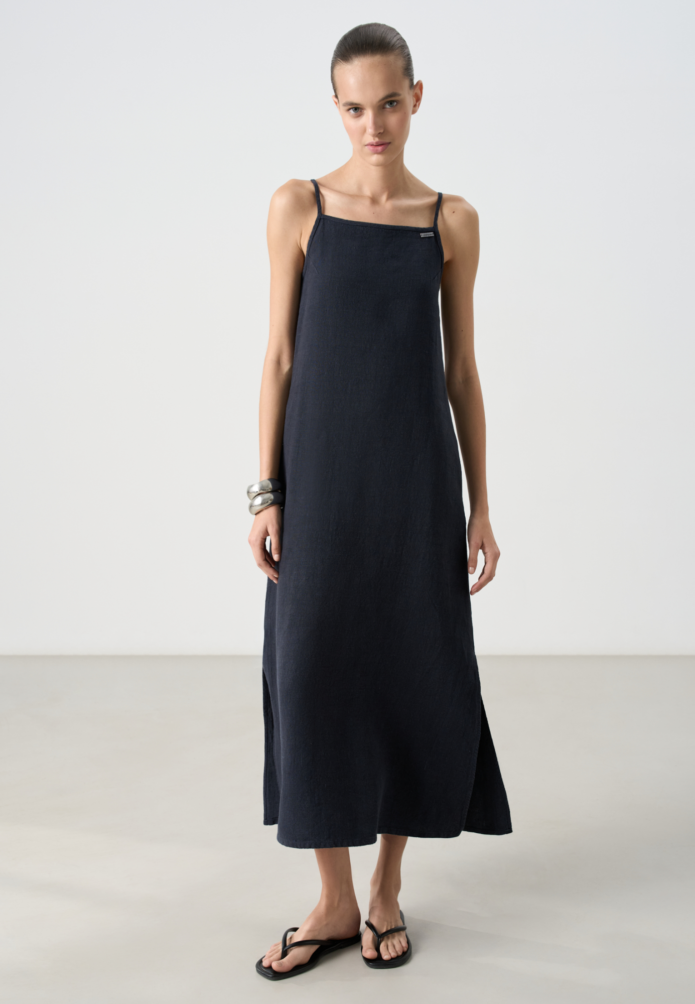 Свободный сарафан длины макси из 100% льна. Модель трапецевидного силуэта, на тонких бретельках и разрезами по бокам. Металический брендированный значок спереди.