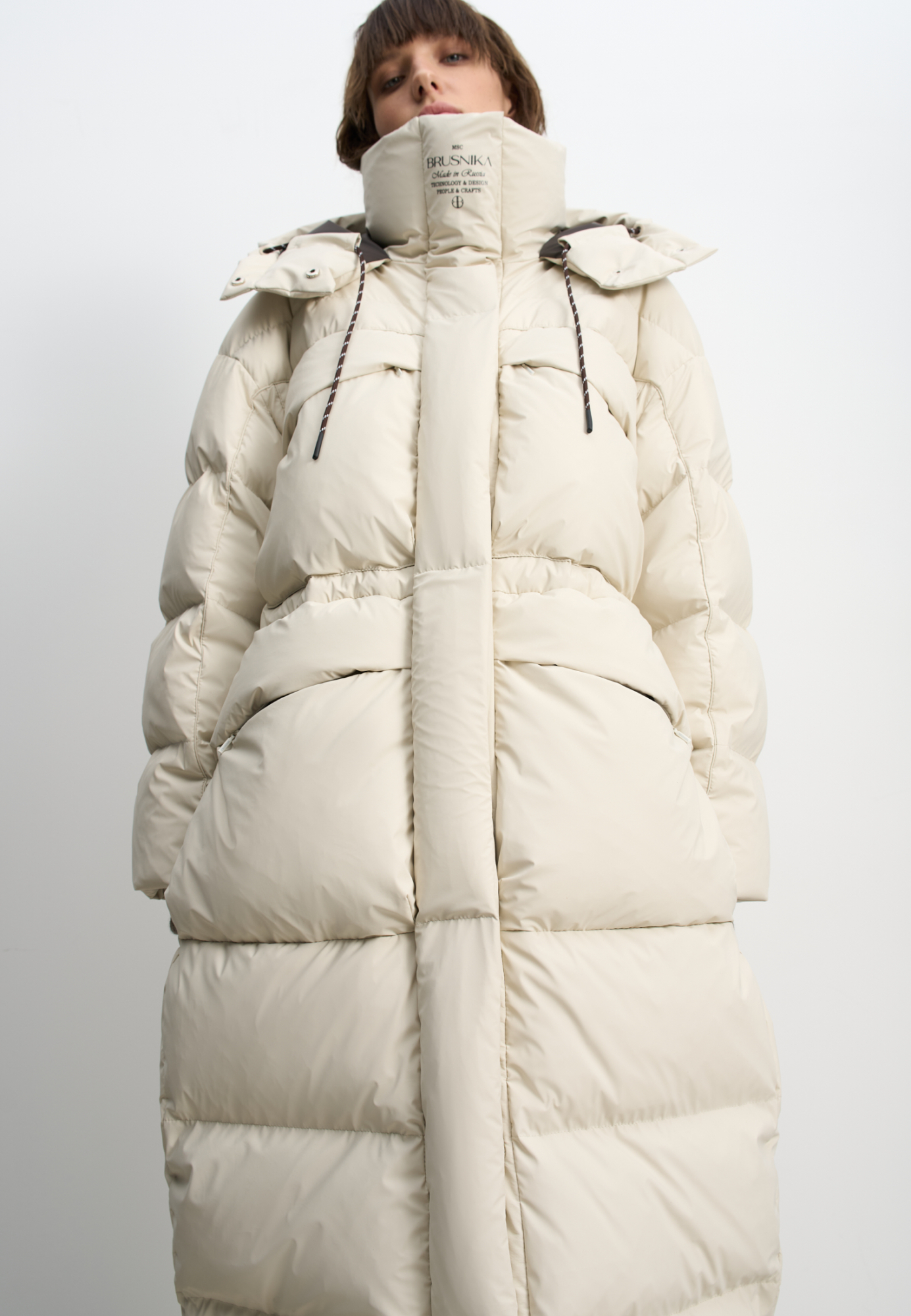 Объемный длинный пуховик. Модель с воротником-стойкой, капюшоном на кулиске и кнопках, большими нагрудными и боковыми карманами на молнии с клапанами и трикотажными манжетами в широкий рубчик. Застежка на молнию по всей длине под планкой на кнопках. Ворот изделия декорирован логотипом бренда.