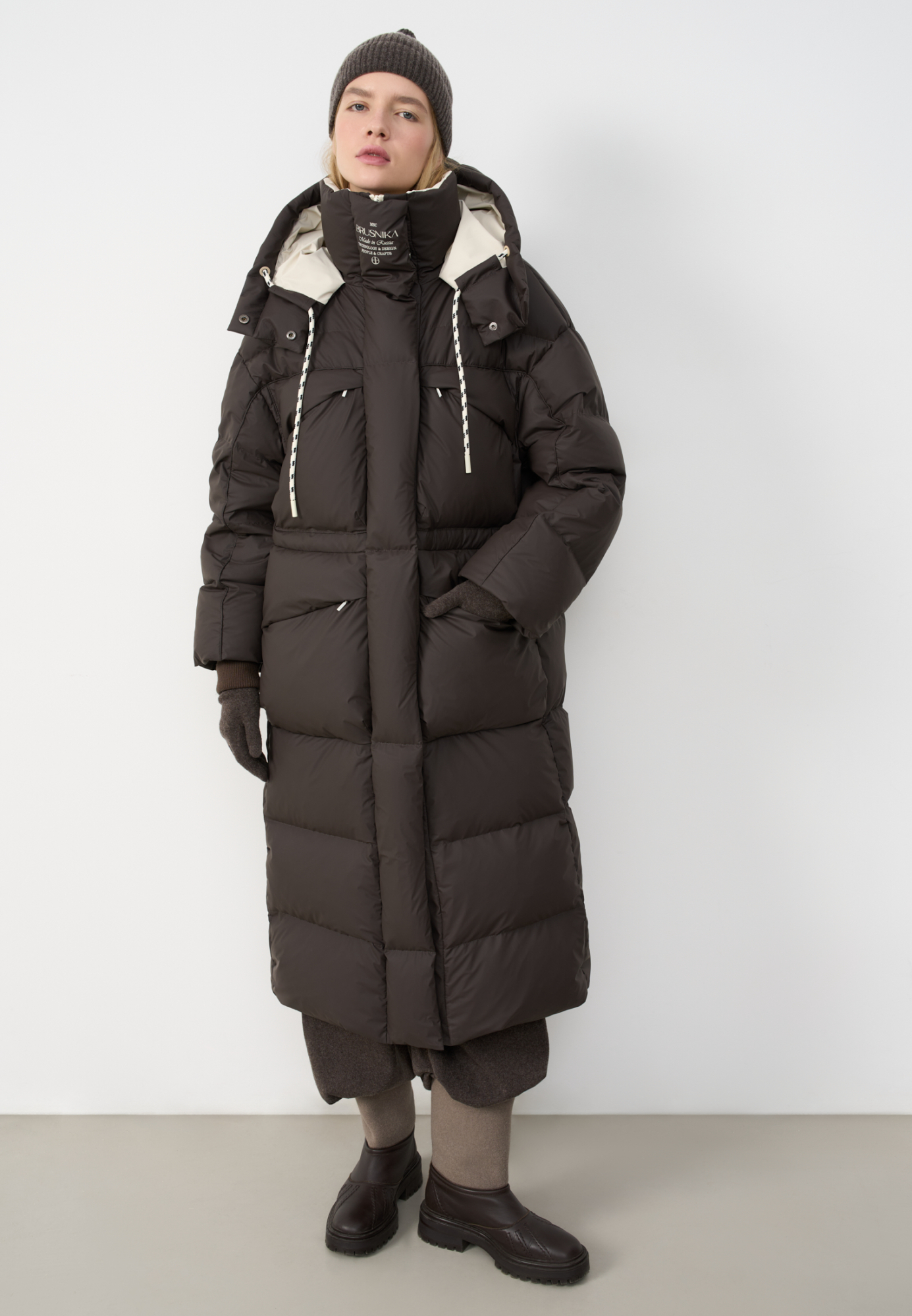 Объемный длинный пуховик. Модель с воротником-стойкой, капюшоном на кулиске и кнопках, большими нагрудными и боковыми карманами на молнии с клапанами и трикотажными манжетами в широкий рубчик. Застежка на молнию по всей длине под планкой на кнопках. Ворот изделия декорирован логотипом бренда.