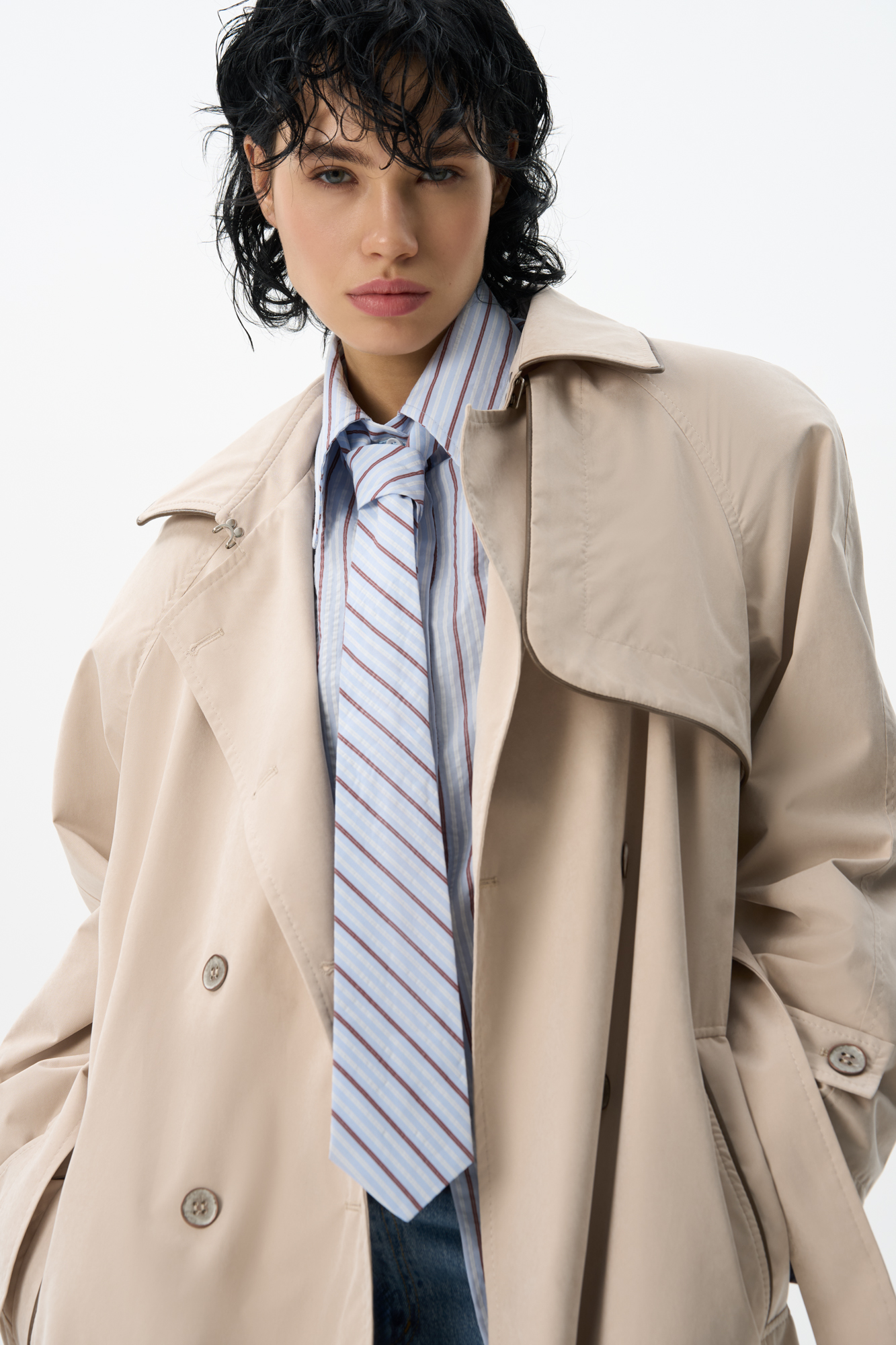 Двубортный длинный тренч прямого кроя. Модель с отложным воротником, декорированным металлической перемычкой, отлетной кокеткой, патами с пуговицами на манжетах, карманами и шлевками на талии для пояса.