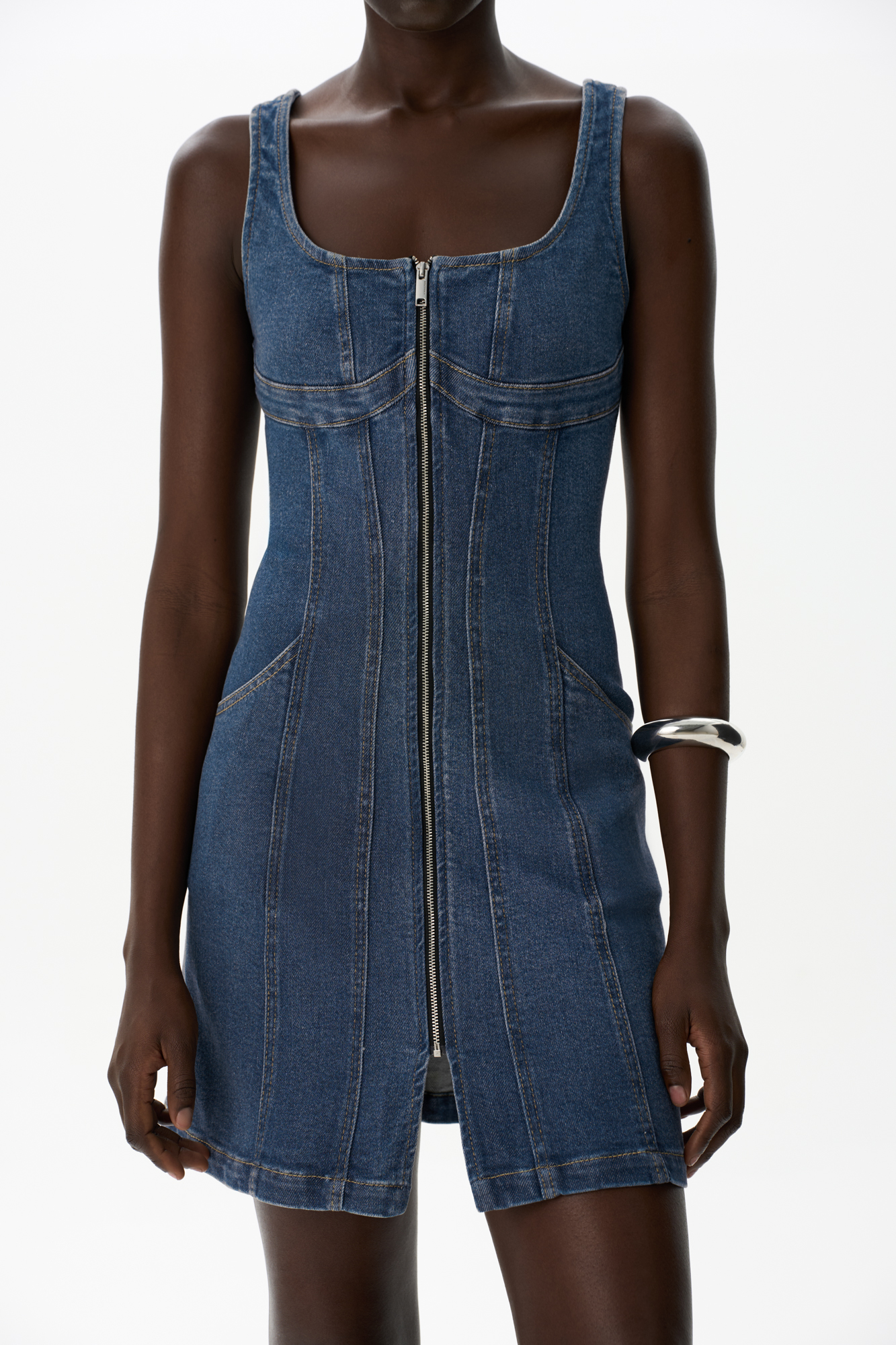 Джинсовое платье длины мини из хлопка. Модель приталенного силуэта на широких бретельках, с двумя боковыми карманами, карманами сзади и застежкой на молнию. Платье имеет отрезной лиф с декоративными швами.