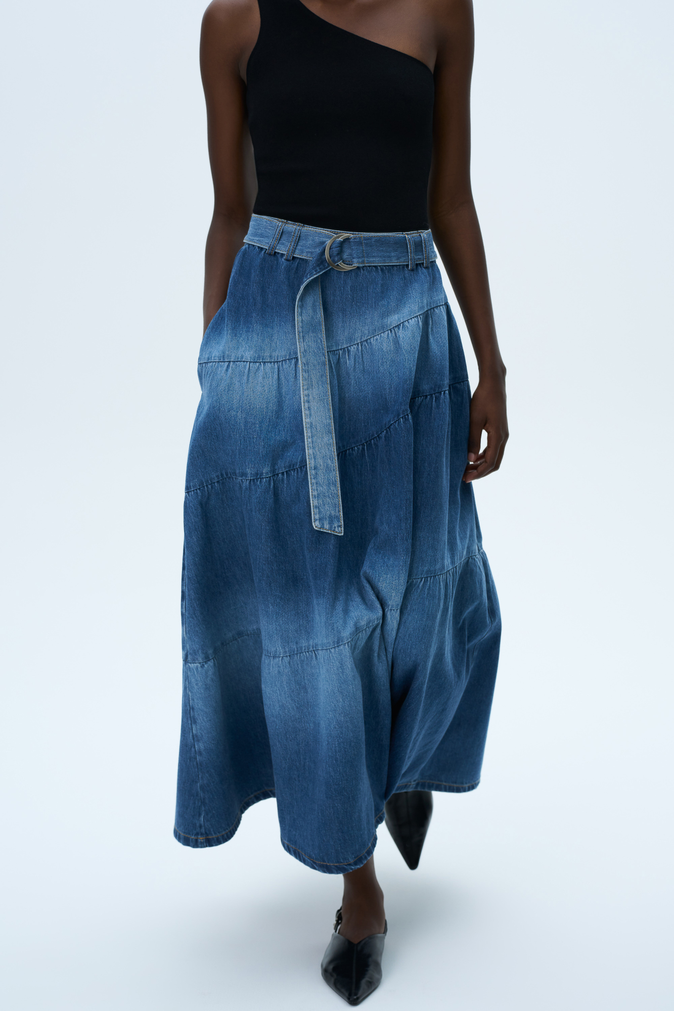 Джинсовая юбка длины миди из 100% хлопка. Модель свободного кроя с застежкой на молнию сзади, шлевками на поясе, ремнем с металлической пряжкой и карманами. Изделие декорировано объемным швом, имитирующим многостойность и ярусы.