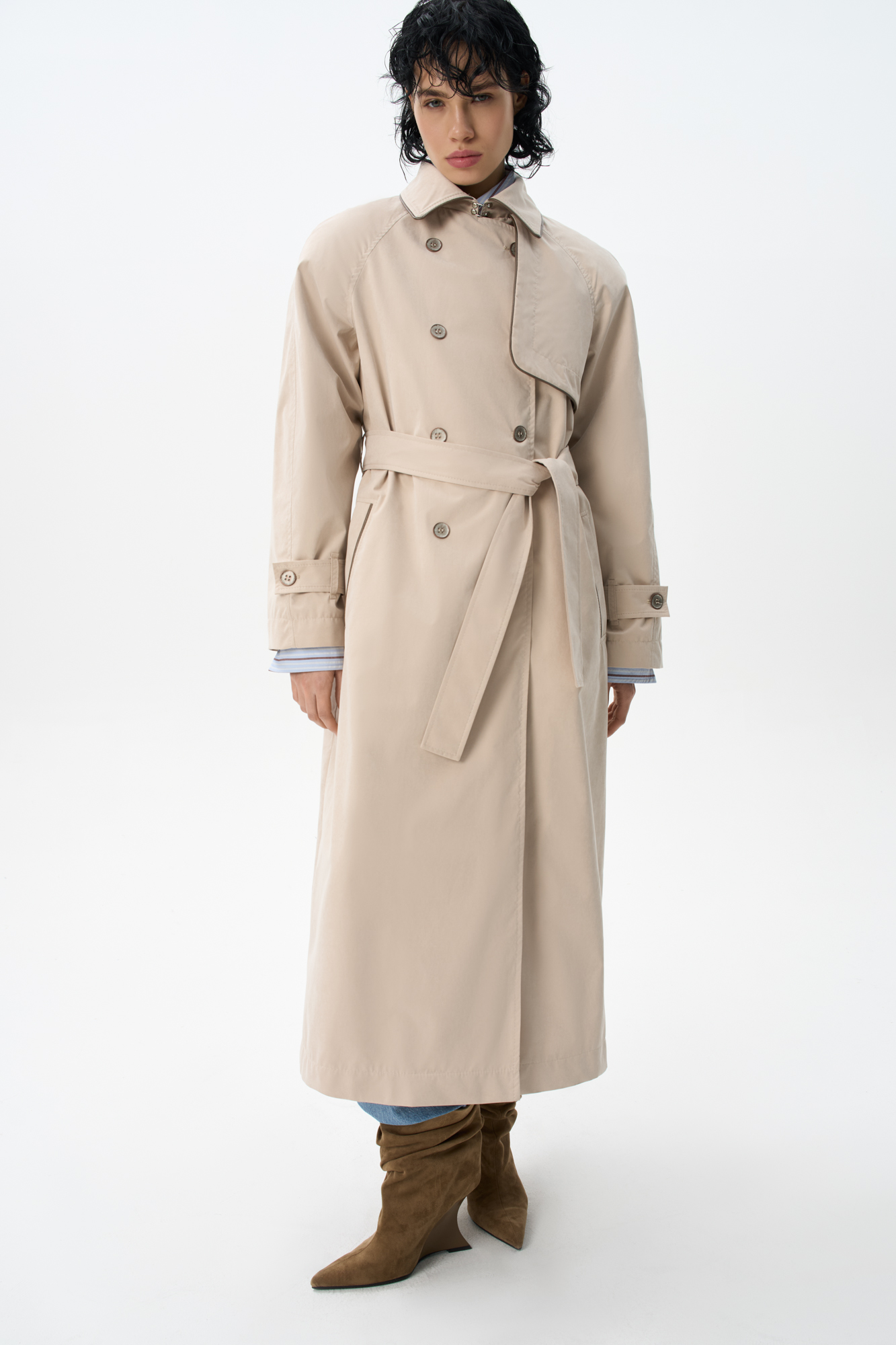 Двубортный длинный тренч прямого кроя. Модель с отложным воротником, декорированным металлической перемычкой, отлетной кокеткой, патами с пуговицами на манжетах, карманами и шлевками на талии для пояса. 
