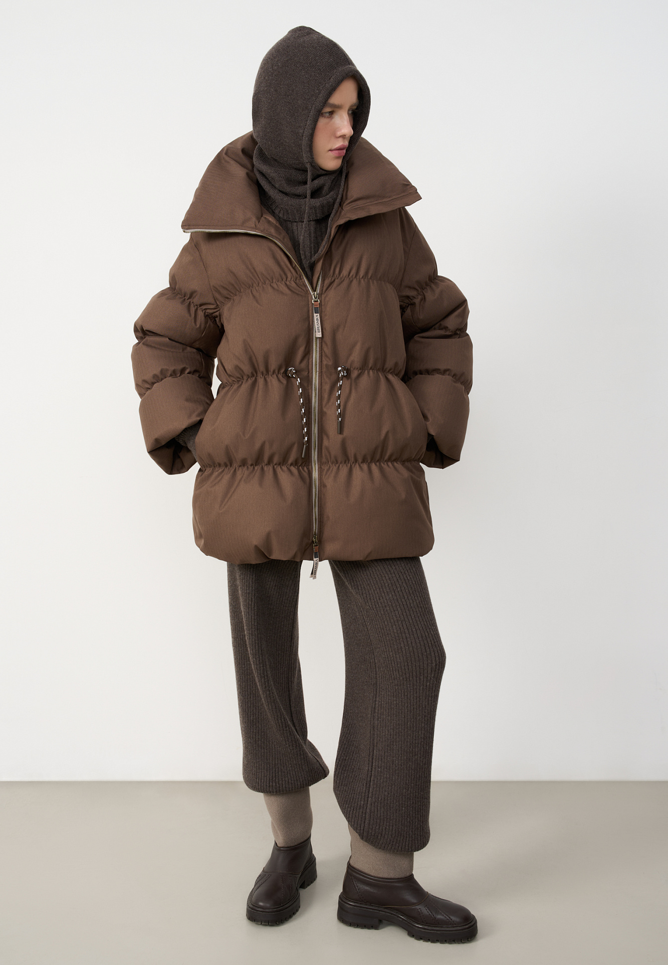 Стеганый объемный пуховик. Модель свободного силуэта, с воротником-стойкой, кулиской на талии и логотипом бренда на спине. Застежка на молнию. 